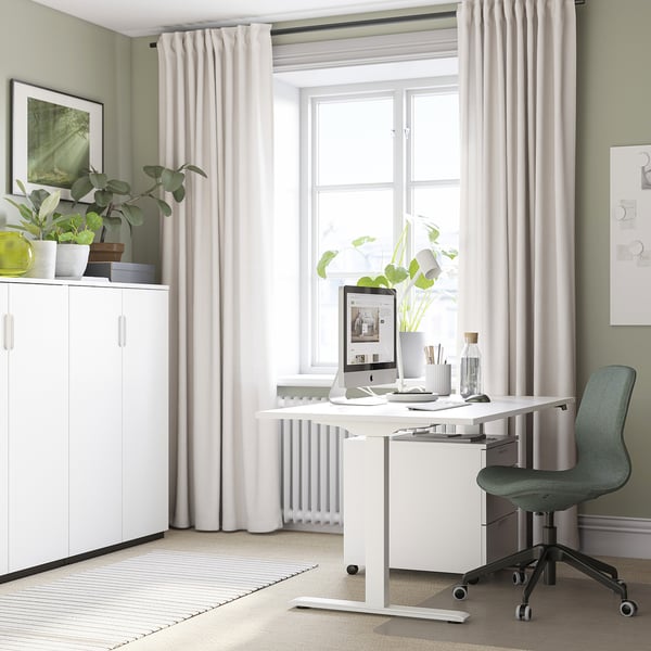 A home office setup with a RODULF white sit-stand desk by a large window. A grey chair, computer, and plants decorate the space.