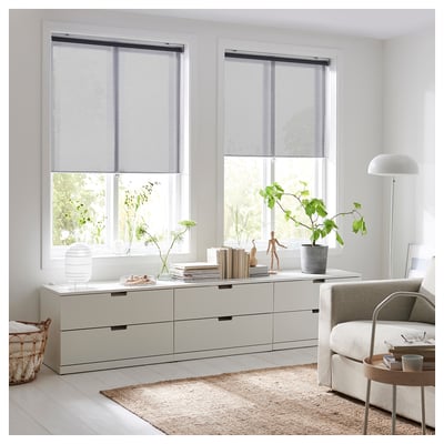 Modern living room with large windows, sheer roller blinds, white furniture, plants, and a jute rug.