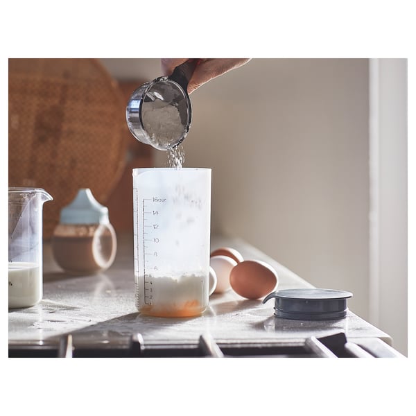 A person shakes powder into a drink or batter using a gradient-marked shaker on a kitchen counter.