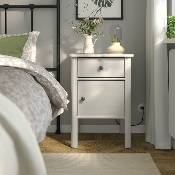White bedside table with 2 drawers, flowers, lamp, between bed & window.