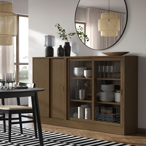 Wooden TONSTAD sideboard with glass doors, mirror, dishes, wicker chair, rug, pendant lamps, minimalist decor.
