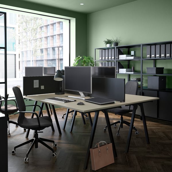 Modern office space with beige TROTTEN desks, black chairs, and large windows. Green walls and natural lighting.