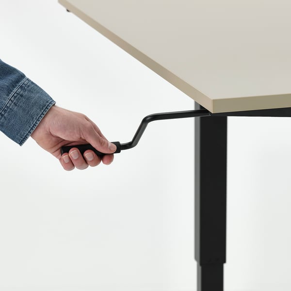 Hand adjusting crank handle of height-adjustable TROTTEN desk, moving between sitting and standing positions.