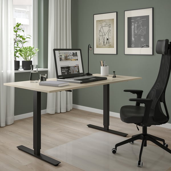 Standing desk with laptop, supplies, plant. Black chair, posters, wood floor, window.