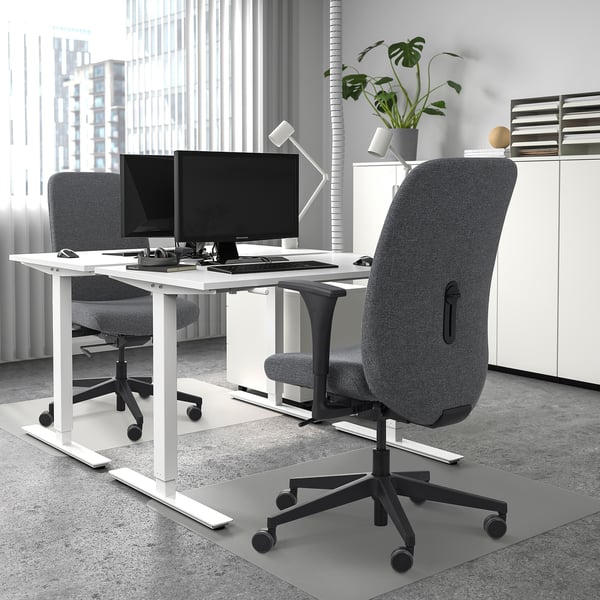 Modern home office with two desks, computers, and a large office chair with greenery.