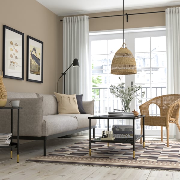Modern living room with neutral tones, black-gold coffee table, grey sofa, wicker chair, artworks, curtains, floor lamp, pillows, rug, balcony view.