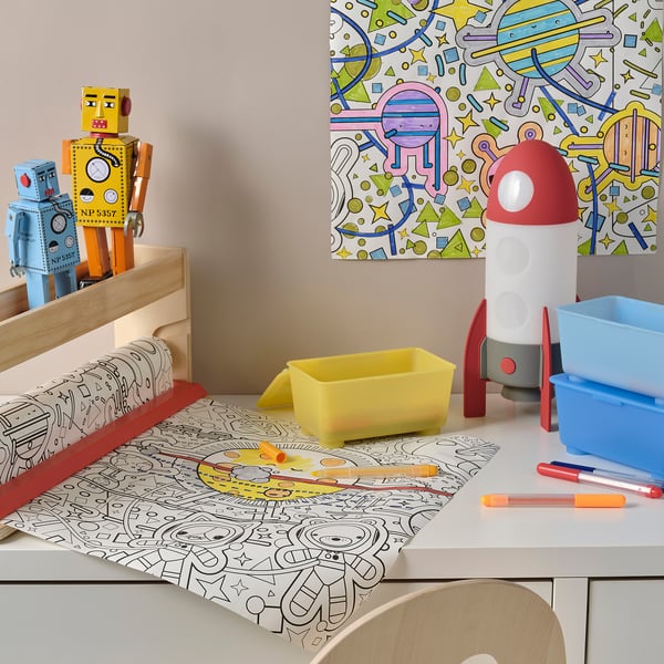 Childs desk with colourful space-themed items: robot toys, colouring mat, pens, and a multi-color led table lamp shaped like a rocket.