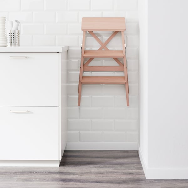 A foldable wooden step ladder leans against a white tiled wall in a kitchen. Its designed for reaching high cabinets and folds to save space.