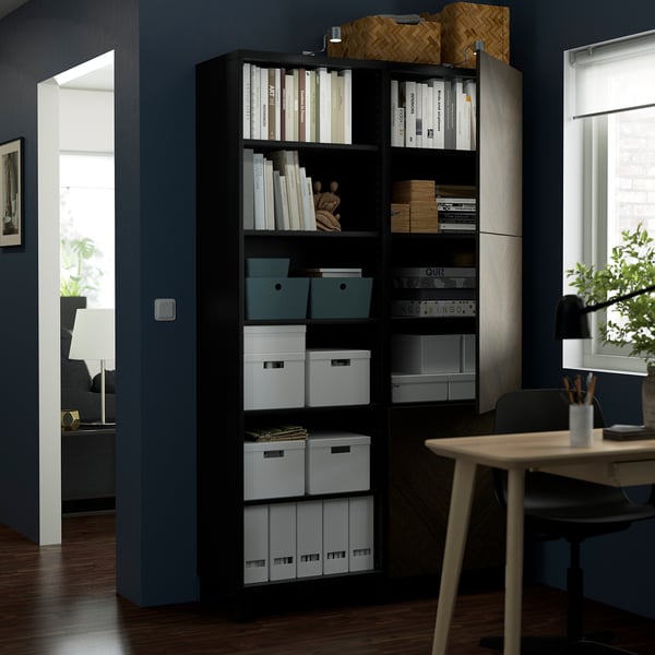 Black BESTÅ storage unit with adjustable shelves, holding books, boxes, and decorative items. Next to a wooden desk with a lamp and chair.