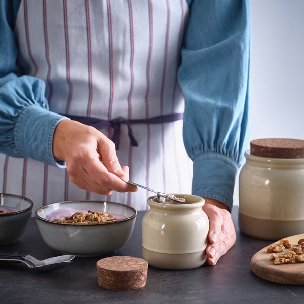 A person holds a jar with a cork lid, scooping contents into a bowl. The jar is part of the BRUGDHAJ series, ideal for storing and serving dry foods.
