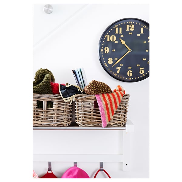 A stylish white shelf with a rattan basket, containing colourful knit items, sits under a large round black clock with gold numbers.