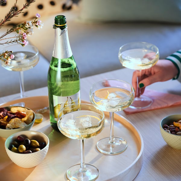 A group of elegant cocktails and appetisers on a table. Four glasses with clear drinks, a green bottle, and bowls of olives, cheeses, and nuts.