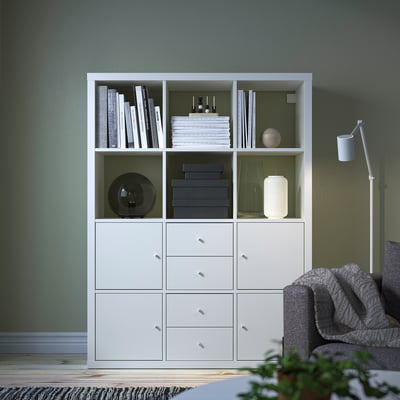 White KALLAX storage unit with shelves and drawers against green wall.