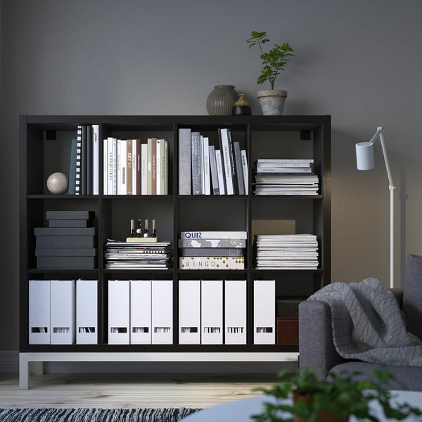 Black KALLAX shelf with books, boxes, blanket, plant, lamp. Modern, organised scene.