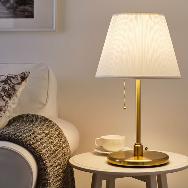 Modern KRYSSMAST lamp with gold base and white shade on side table, beside teacup. Compact size next to armchair with blanket.