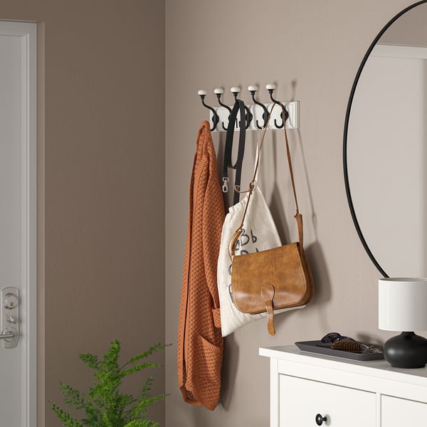 Hook rack, wall-mounted, white with black hooks, holds bags, jackets, small items. Situated near door, mirror, plant.