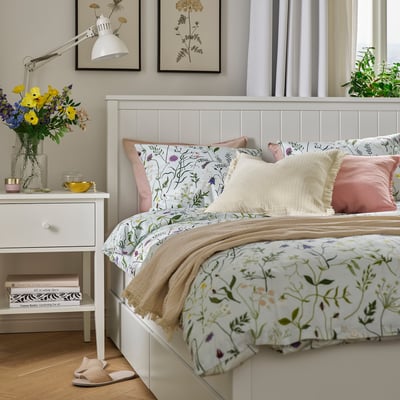 Cosy bedroom with MATTOXBÄR pale blue floral duvet. White bed with pink pillows, beige blanket. Beside table has flowers, lamp, books.