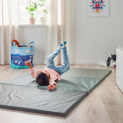 Baby playing on a soft, foldable PASSBIT gym mat, practising somersaults indoors. Nearby, a toy bin and stuffed animals are visible.