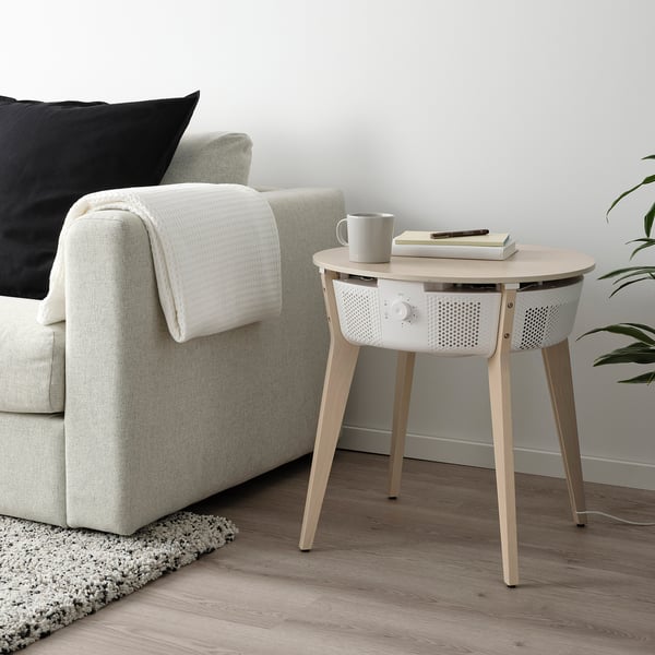 A white STARKVIND air purifier side table beside a grey couch. Round, wooden, holds a mug and book. Ideal for small spaces.