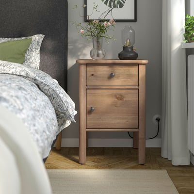 Wooden STOREMOLLA bedside table by bed with lamp and flowers.