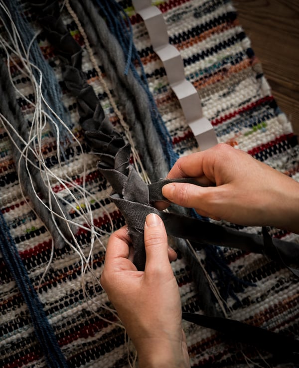 A person is crafting a TÅNUM rug from black cotton strips. The rug intricate patterns on a multi-colored woven base.