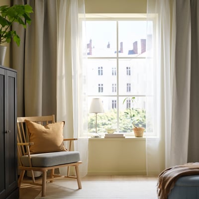 Bright room with large white TERESIA curtains. Chair, lamp, and plant nearby. Cosy, airy feel enhanced by floor-length curtains.