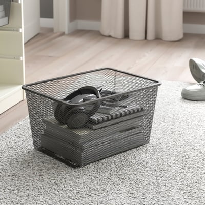 Mesh storage basket on carpet, containing headphones, near a wooden floor and white curtains.