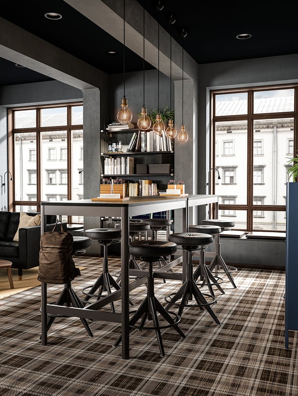 Modern office space with high tables, black stools, large windows, shelves with books, and hanging lights.