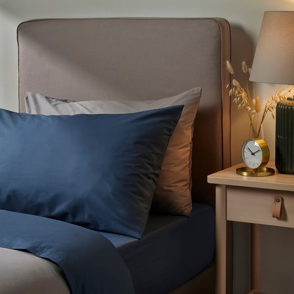 Dark blue pillowcase on bed. Soft cotton-lyocell blend, square shape. Grey headboard, bedside table with vase and clock.