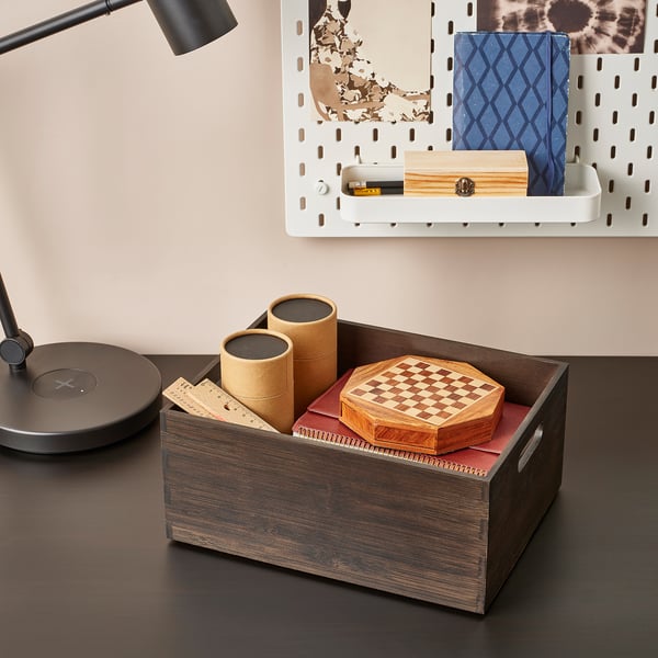 Wooden box on desk holding chess set, cylindrical containers, and ruler. Desk lamp and wall-mounted storage in background.