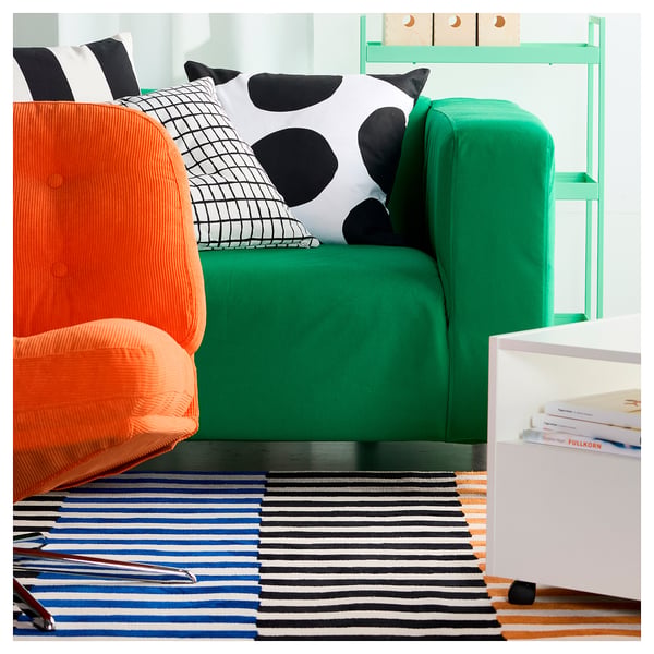 Bright orange and green chairs with black and white pillows on a striped rug, next to a white table with colourful books.