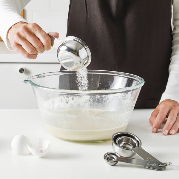 A person pours sugar from a measuring spoon into a bowl; measuring spoons used.