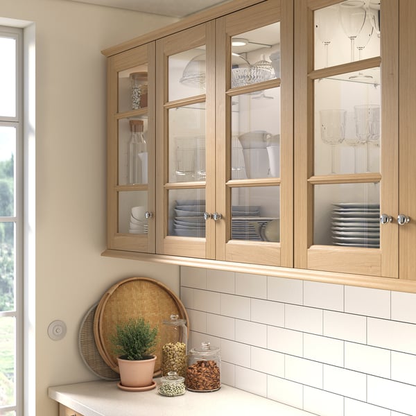 Kitchen cabinets with glass doors showing plates, glasses, jars. Brown oak finish with wicker tray and plant on counter.