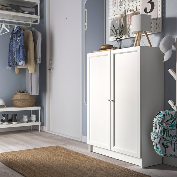 White BILLY bookcase in entryway with closed doors, next to coat rack, on beige rug, with basket and backpack nearby, light blue wall with pegboard.