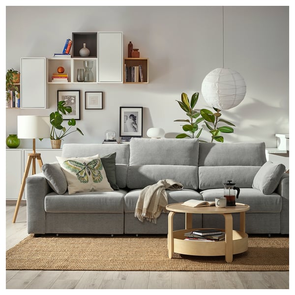 Modern living room with grey ESKILSTUNA sofa, wooden coffee table, plants, and wall shelves displaying books and decorations.