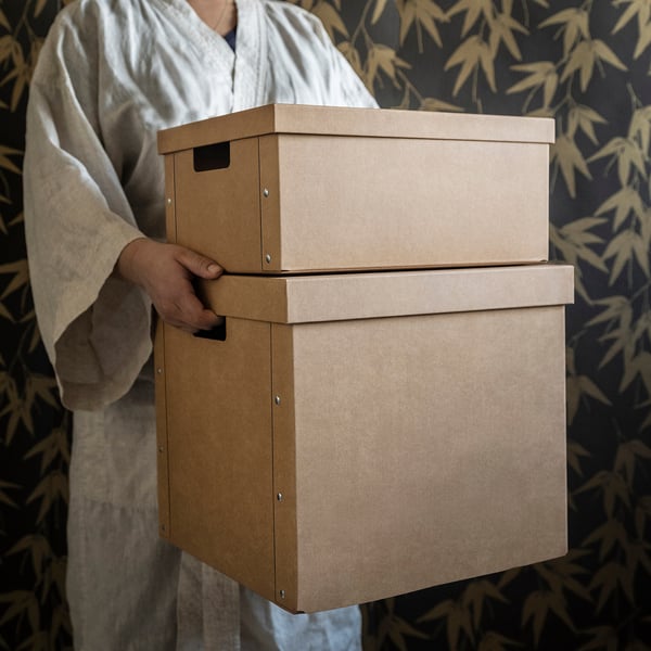 Person in white robe holding stack of brown storage boxes with handles.