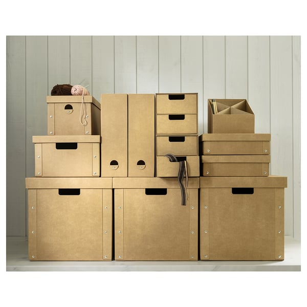 A stack of GÄRDESGÅRD cardboard storage boxes in various sizes and shapes, brown colour, placed on a shelf against a beige wooden wall.