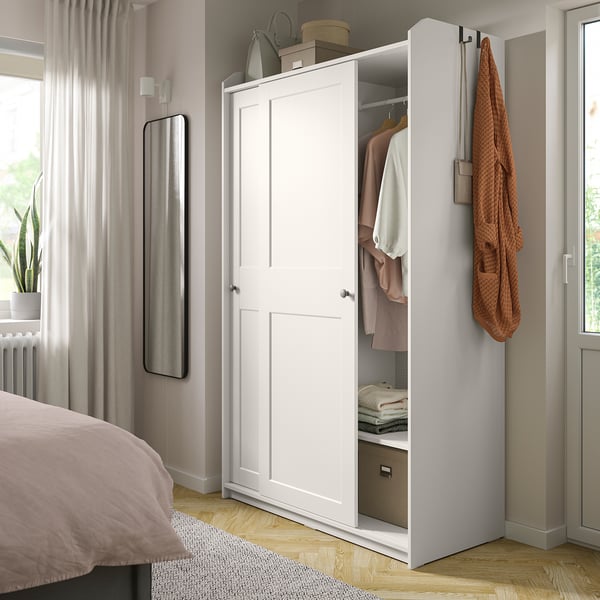 A HAUGA white wardrobe with sliding doors in a bedroom. One door is open, revealing clothes and storage boxes. A mirror and plant are nearby.