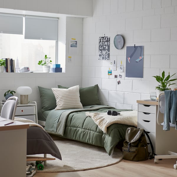 Bedroom with green HEDFRYLE comforter, desk, chair, plant on table, white brick walls.