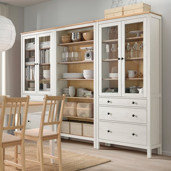 HEMNES white storage unit with glass doors and drawers, holding dishes and glasses, next to wooden chairs.