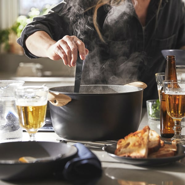Person cooking with large pan, steam rising, surrounded by drinks and slices of bread.
