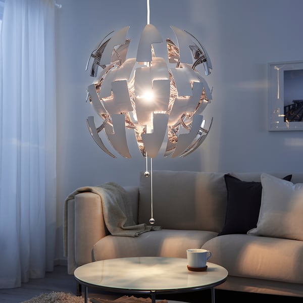 Modern living room with IKEA PS 2014 chrome pendant lamp, intricate design, hanging above a cosy grey sofa and a round coffee table with a mug.