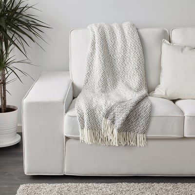 White couch with grey throw blanket draped over it, featuring fringes and a textured pattern.