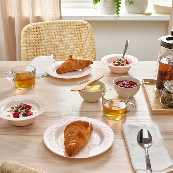 Wooden table set with white dinnerware. Includes bowls of fruit, croissants, cups of tea, and a glass pitcher. Dinnerware has a patterned rim.