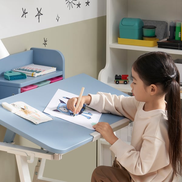 A young girl sits at a blue, adjustable-height PIPLÄRKA desk, drawing on paper; the tiltable desk surface and hooks are visible.