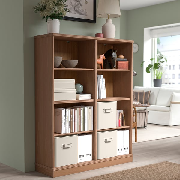 Light wood PLOGFÅRA storage unit with five sections. Stores books & decor. Beige bins & bowl, horse figurine. Tall, fits near seating area.