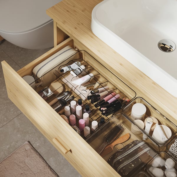 Organised light wood bathroom drawer with clear acrylic dividers storing makeup and hair tools.
