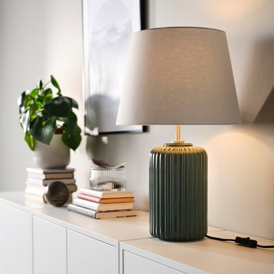 SNÖBYAR lamp: dark green ceramic base, beige cloth shade, tall on table beside books, plant, and glass.