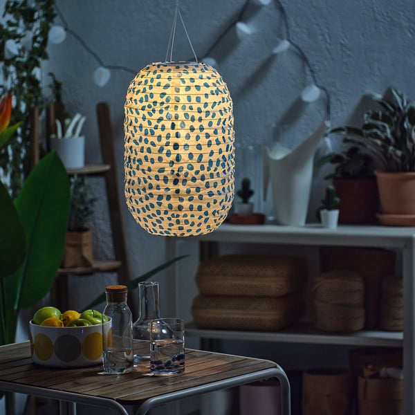 A lit solar-powered lantern with blue dots hangs above a table with a bowl of lemons. The lantern lights up automatically at dusk.