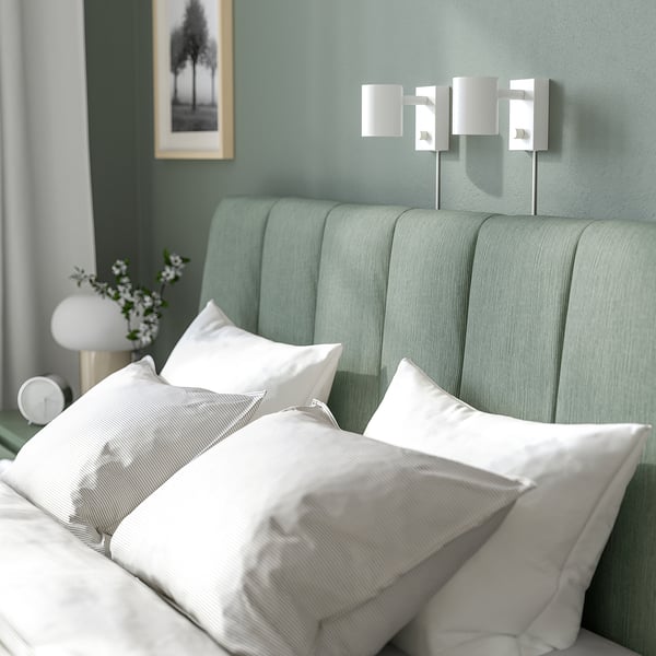 Modern bed with grey-green headboard, white pillows, flowers, and wall lamps.